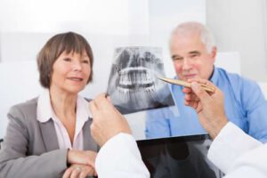 A dentist shows a dental X-ray to an older woman and man in an office. The woman is wearing a gray blazer, and the man is in a blue shirt. The dentist holds a pen, pointing at the X-ray details.