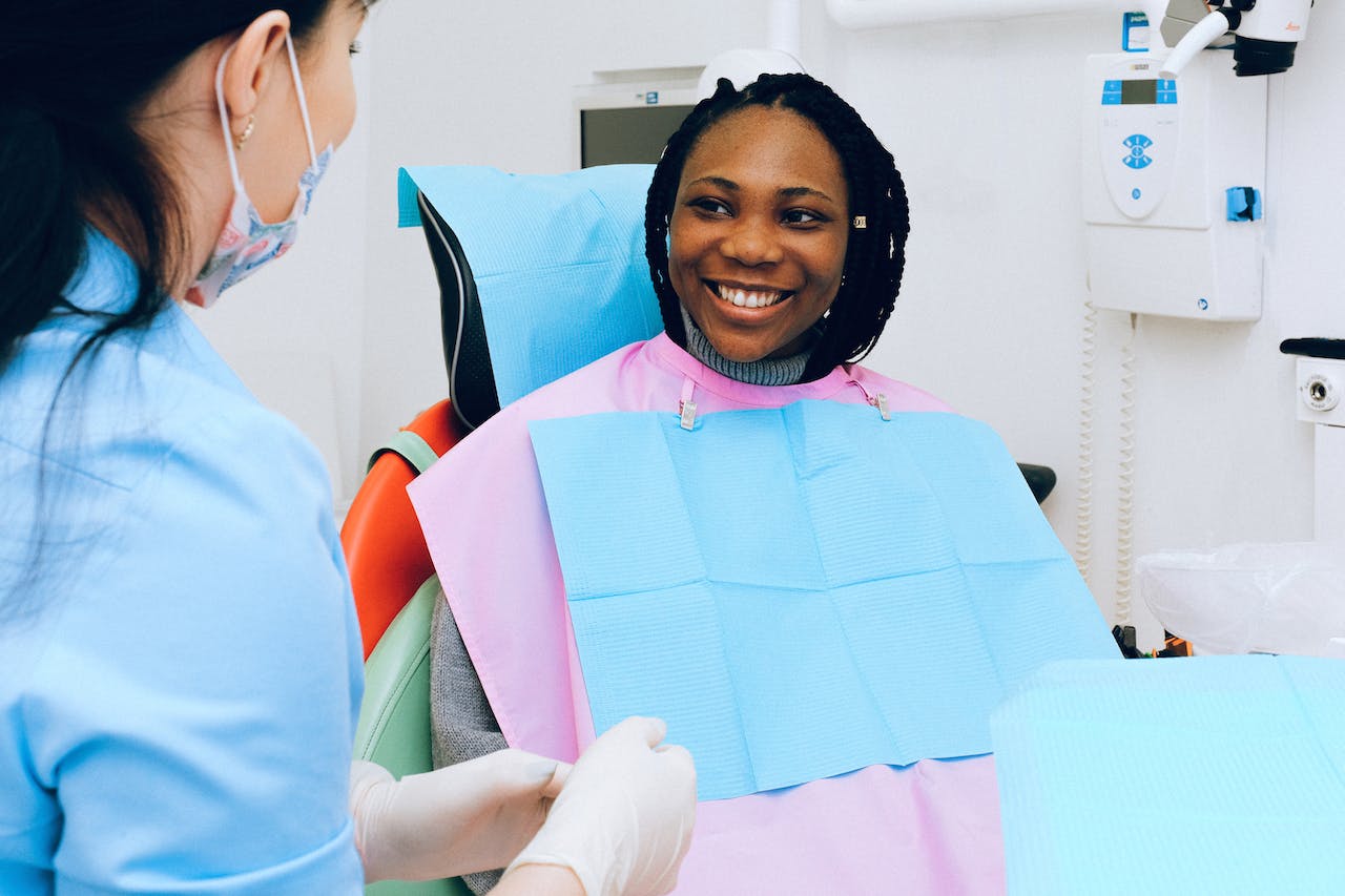 A patient wearing a dental bib smiles at a dentist in a clinic. The dentist is wearing gloves and a mask, facing the patient. The setting appears clean and professional, with dental equipment visible in the background.