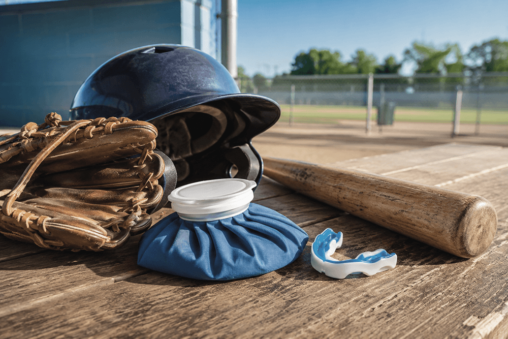 Baseball gear on a sunlit bench next to an ice pack and mouth guard.